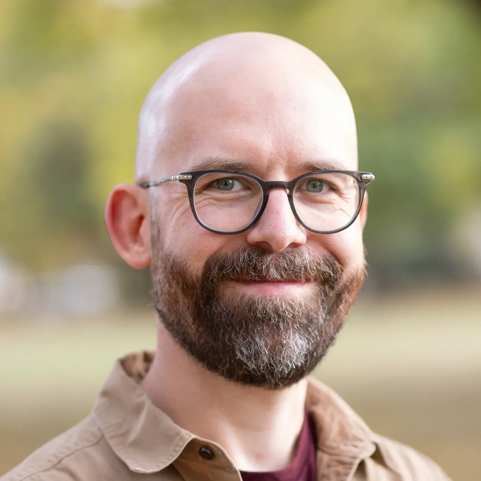 Me, Josh Collinsworth, a bald white guy with a beard and glasses, smiling against a blurry autumn background in a button-up shirt.